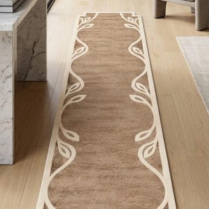 May include: A long, rectangular runner rug featuring a solid tan center and a cream-colored border. The border is decorated with a continuous, symmetrical vine and leaf pattern that runs along both sides of the rug, framing the tan interior.