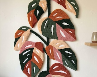 Puede incluir: Un tapiz de pared con técnica de tufting que representa una planta monstera con hojas variegadas en tonos verde oscuro, naranja óxido, rosa claro y crema. La pieza textil presenta un tallo central verde oscuro y el texto MAVIS HA. abajo a la derecha.