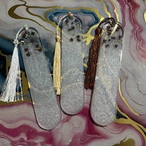 May include: Three rectangular resin bookmarks with rounded ends, each filled with silver glitter and small dark heart-shaped accents. Each bookmark features a decorative tassel attached to the top: one white, one gold, and one dark brown.