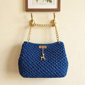 May include: A deep blue crocheted handbag featuring a gold-toned metal chain strap and a rectangular gold-toned clasp. A small gold-toned deer charm hangs from the center of the clasp. The bag is suspended from two gold-toned wall hooks.