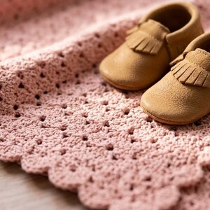 May include: A pair of tan moccasins with fringe detailing placed on a soft, light pink crocheted blanket. The blanket features a repeating openwork pattern with a scalloped edge, providing a textured base for the two small slip-on shoes.
