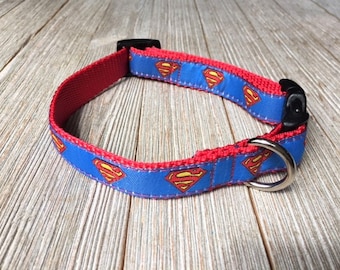 May include: A red and blue dog collar with a Superman logo pattern. The collar has a black plastic buckle and a silver metal D-ring.