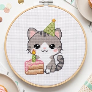 May include: A cross-stitch embroidery in a round wooden hoop featuring a gray and white tabby cat wearing a green party hat. The cat sits next to a pink frosted birthday cake with a single lit green candle, all stitched on white fabric.