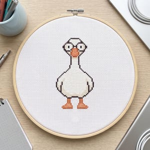 May include: A circular wooden embroidery hoop containing a white Aida fabric piece with a cross-stitch design of a white goose. The goose has a bright orange beak and feet, black outlines, and large circular eyes with black pupils centered on its head.
