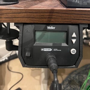 Op de afbeelding: Een zwarte Weller soldeerstation-besturingseenheid gemonteerd onder een houten bureau. Het apparaat is voorzien van een digitaal display, een aan-uitknop, navigatiepijlen, een menuknop en een label met 80 YEARS LIMITED BLACK EDITION.