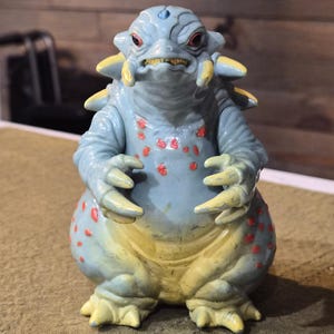 May include: A blue monster figurine with a rounded body, yellow accents on the feet and spikes, and red spots on the chest and sides. The creature has a wide head with red eyes, a small blue gem on the forehead, and sharp claws on its hands.