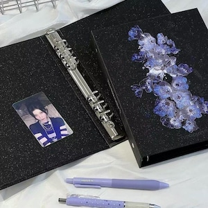 Op de afbeelding: Een zwarte glittermap met een cluster van blauwe bloemillustraties op de kaft, een zilverkleurig metalen mechanisme met zes ringen en een rechthoekige fotokaart van een persoon in een gestreept shirt. Twee lichtpaarse pennen liggen onder de map.