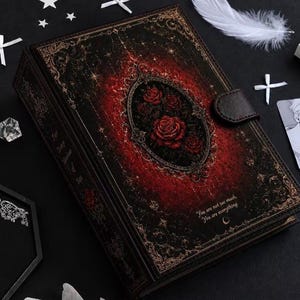 Puede incluir: Un diario rectangular negro con cierre magnético, que presenta un marco ovalado central con rosas rojas. La cubierta tiene bordes de filigrana dorada, motivos de estrellas y el texto: You are not too much, You are everything.
