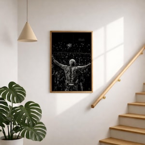 Op de afbeelding: Een rechthoekige ingelijste wandkunstprint met een zwart-witfoto van een vechter die met uitgestrekte armen in een volle arena staat. De vechter heeft een zichtbare rugtattoo en draagt een broek met de merknaam Venum erop gedrukt.