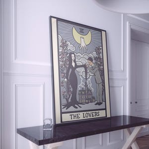 May include: A framed rectangular wall art print featuring a tarot card design titled THE LOVERS. The illustration depicts a woman in a black dress and a man in a pinstripe suit standing before a house, with a large yellow moon and hand above.