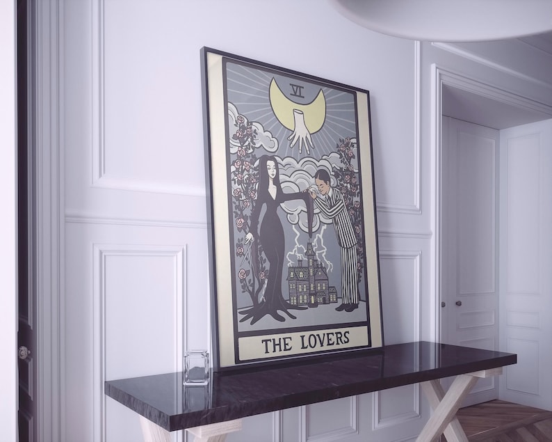 May include: A framed rectangular wall art print featuring a tarot card design titled THE LOVERS. The illustration depicts a woman in a black dress and a man in a pinstripe suit standing before a house, with a large yellow moon and hand above.