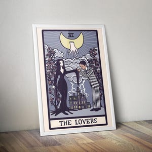 May include: A framed art print featuring a tarot card design titled THE LOVERS. The illustration depicts a person in a long black dress and a person in a striped suit standing before a house, with a disembodied hand and a crescent moon above.