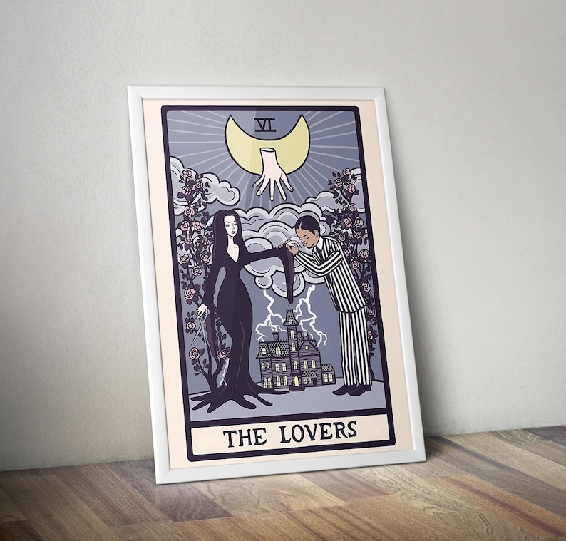 May include: A framed art print featuring a tarot card design titled THE LOVERS. The illustration depicts a person in a long black dress and a person in a striped suit standing before a house, with a disembodied hand and a crescent moon above.