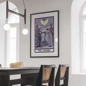 May include: A framed wall art print featuring a tarot card illustration titled THE LOVERS. The artwork depicts a couple in a gothic style, a large house, and a crescent moon with a hand above. The color palette includes shades of purple, black, and cream.