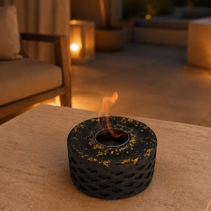 May include: A circular tabletop fire pit with a textured dark grey concrete exterior featuring gold-coloured flecks and a central metal burner insert containing a small, flickering orange flame, resting on a light beige stone surface.
