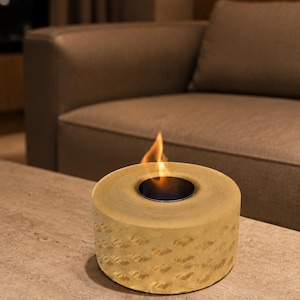 May include: A cylindrical tabletop fire pit with a textured, light beige stone-like exterior and a central black metal burner insert. A small, flickering orange flame rises from the centre of the circular opening, casting a warm glow.