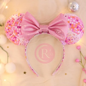 Puede incluir: Una diadema con dos orejas circulares cubiertas con un patrón de bola de discoteca en rosa, naranja y blanco. Un lazo grande, brillante y de color rosa claro con un acabado de purpurina sutil está centrado entre las orejas. La diadema tiene un logo.