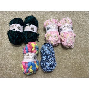 May include: Six skeins of Gala Yarns Mixed Fiber yarn. Two are dark green, two are a mix of pink, cream, and yellow, one is a multi-colored blend of pink, blue, and yellow, and one is a dark blue and white speckled yarn. Each has a white label.
