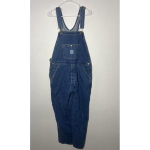 May include: A pair of dark blue denim overalls featuring adjustable shoulder straps with metal buckles, a front bib pocket with a small rectangular brand patch, side button closures, and two deep hip pockets. The garment has a classic straight-leg fit.