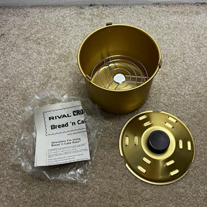 May include: A gold-colored metal baking canister with a removable wire rack inside, a matching gold lid with a black knob, and a plastic-wrapped instruction manual labeled Rival Crock-Pot Bread 'n Cake Bake with usage directions.