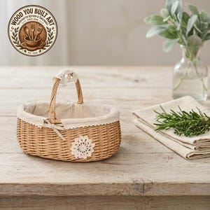 May include: An oval woven wicker basket with a single arched handle, featuring a beige fabric liner with a lace trim edge, a small fabric bow, and a white crocheted flower appliqué on the front exterior, presented with a logo in the corner.