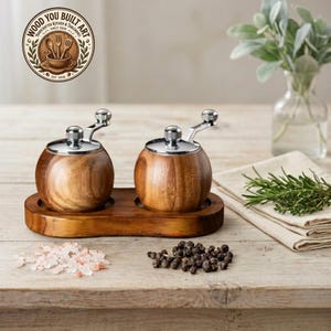 Op de afbeelding: Twee ronde houten kruidenmolens met zilverkleurige metalen zwengels staan op een bijpassende houten voet. De set bevat een cirkelvormig logo in de linkerbovenhoek met de tekst Wood You Built Art, Handcrafted Kitchen & Tablewares.