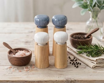 May include: Two wooden salt and pepper grinders with ribbed blue and white tops, accompanied by two small dark wooden bowls containing pink salt and black peppercorns, each with a small wooden spoon, arranged on a light wooden surface.