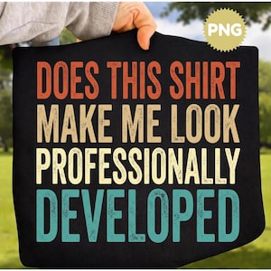 May include: A black t-shirt featuring the text DOES THIS SHIRT MAKE ME LOOK PROFESSIONALLY DEVELOPED in a retro-style font. The words are printed in three distinct colors: orange for the first line, beige for the middle, and teal for the bottom.