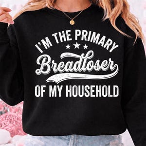 Könnte beinhalten: Ein schwarzes Langarm-Sweatshirt mit weißem Text, der I'M THE PRIMARY Breadloser OF MY HOUSEHOLD lautet, mit drei kleinen Sternen über dem Wort Breadloser. Das Design ist in einer fetten, abgenutzten weißen Schriftart gedruckt.