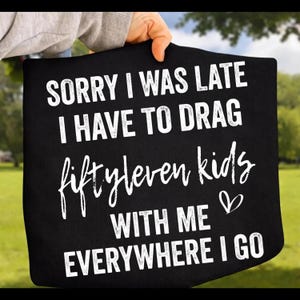 May include: A black t-shirt featuring white text that reads: SORRY I WAS LATE I HAVE TO DRAG fiftyleven kids WITH ME EVERYWHERE I GO. The word fiftyleven is written in a cursive script font, and a small white heart icon is positioned to the right.