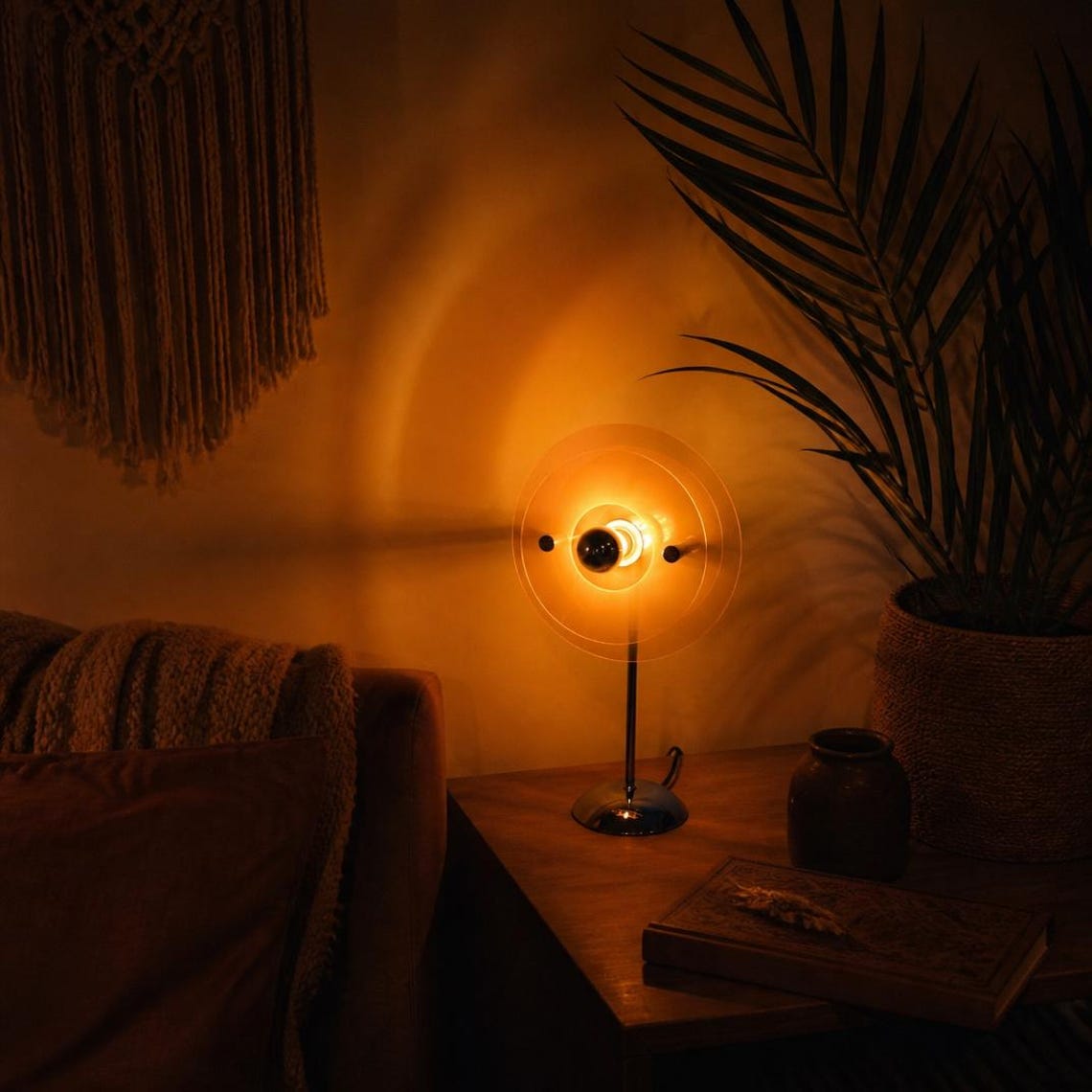 May include: A table lamp featuring a thin, metallic stand and a circular, translucent amber-colored glass shade. The lamp emits a warm, glowing light from a central bulb, casting a soft, circular illumination pattern against the surrounding wall.