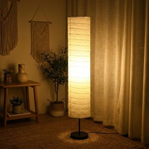 May include: A tall, cylindrical floor lamp featuring a textured, off-white paper shade with horizontal ribbed detailing. The lamp is supported by a thin, black metal pole and a circular black base, emitting a warm, soft glow from within.
