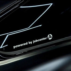 Könnte beinhalten: Ein weißer Vinyl-Aufkleber auf einer dunklen, reflektierenden Oberfläche. Der Aufkleber zeigt den Text "powered by jobcenter" in einer serifenlosen Schrift, begleitet von einem kreisförmigen Logo mit einem stilisierten Buchstaben A in Weiß.