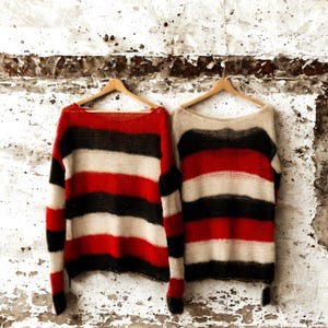 Op de afbeelding: Twee gestreepte truien hangen aan houten kleerhangers tegen een verweerde witte muur. Eén trui heeft rode, zwarte en crèmekleurige horizontale strepen. De andere trui heeft vergelijkbare strepen, waarbij crème het rood vervangt. Beide truien hebben lange mouwen.