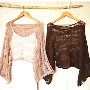 May include: Two loose-knit, long-sleeved cropped sweaters hanging on wooden hangers. One sweater is a light dusty pink, layered over a white tank top, and the other is a solid dark brown. Both garments feature a wide, open-weave knit texture.