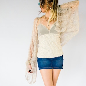 Lightweight beige knit shrug for summer layering, shown styled with jeans and layered necklaces. This airy, hand-knit piece features relaxed drape and extra-long sleeves, perfect for festival wear, beach cover-ups, or everyday bohemian style.
