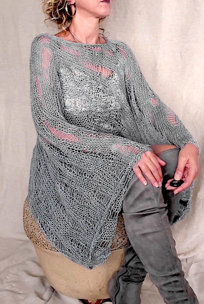 Op de afbeelding: Een grijze, open geweven gebreide tuniek met lange mouwen en een boothals. De tuniek heeft distressed details en wordt gecombineerd met grijze over-de-knie laarzen. De algehele stijl is bohemien.