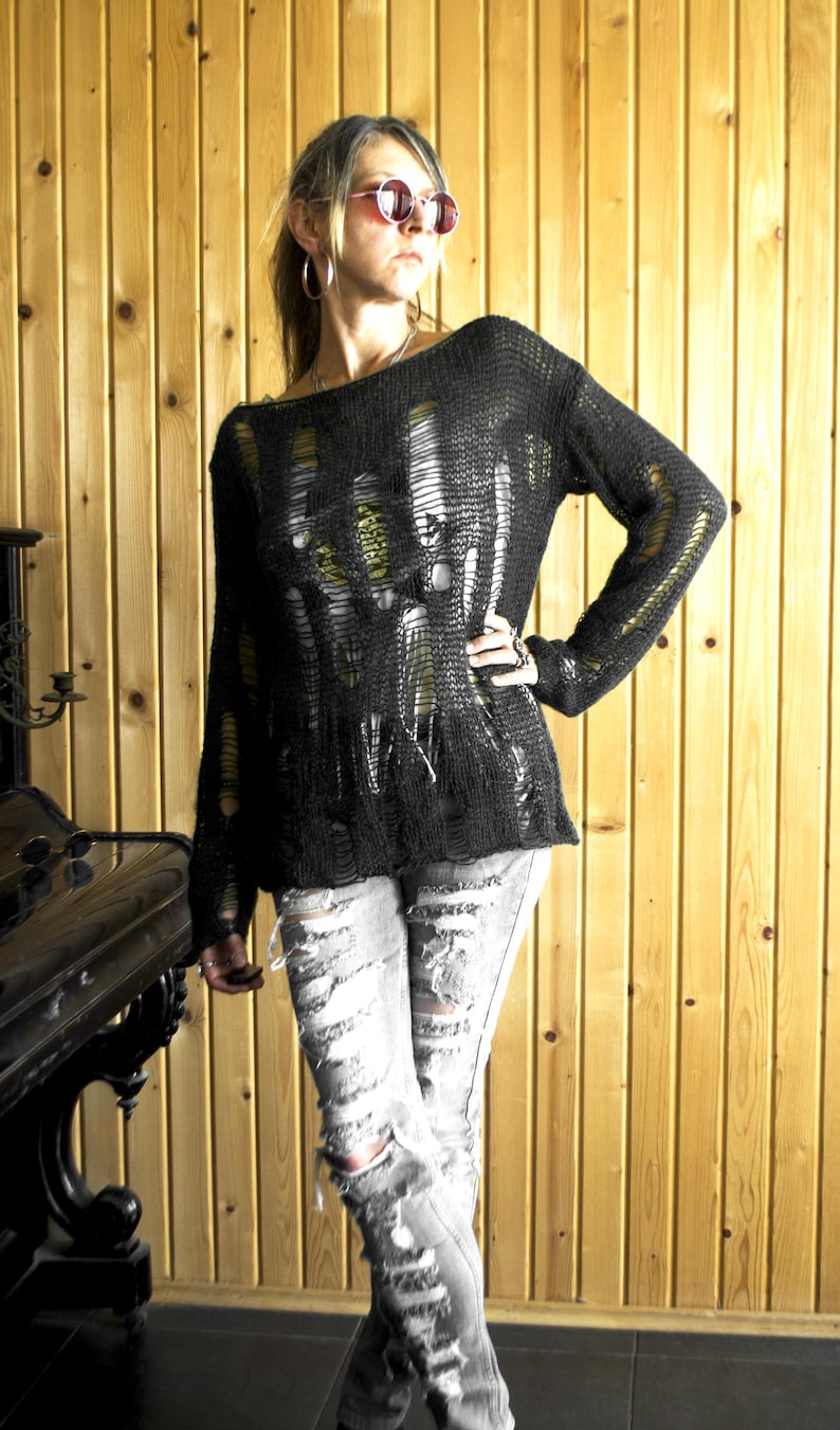 A black, distressed knit sweater with long sleeves and a shredded design. The sweater is paired with ripped gray jeans. The model is wearing pink sunglasses and hoop earrings. The background is a wooden wall.