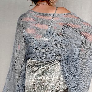 Op de afbeelding: Een lichtblauwe, open geweven gebreide sjaal met lange mouwen. De sjaal is over een rok met patroon gedrapeerd. Het breisel heeft een los, luchtig ontwerp. De rok heeft een paisley patroon in grijstinten en wit.