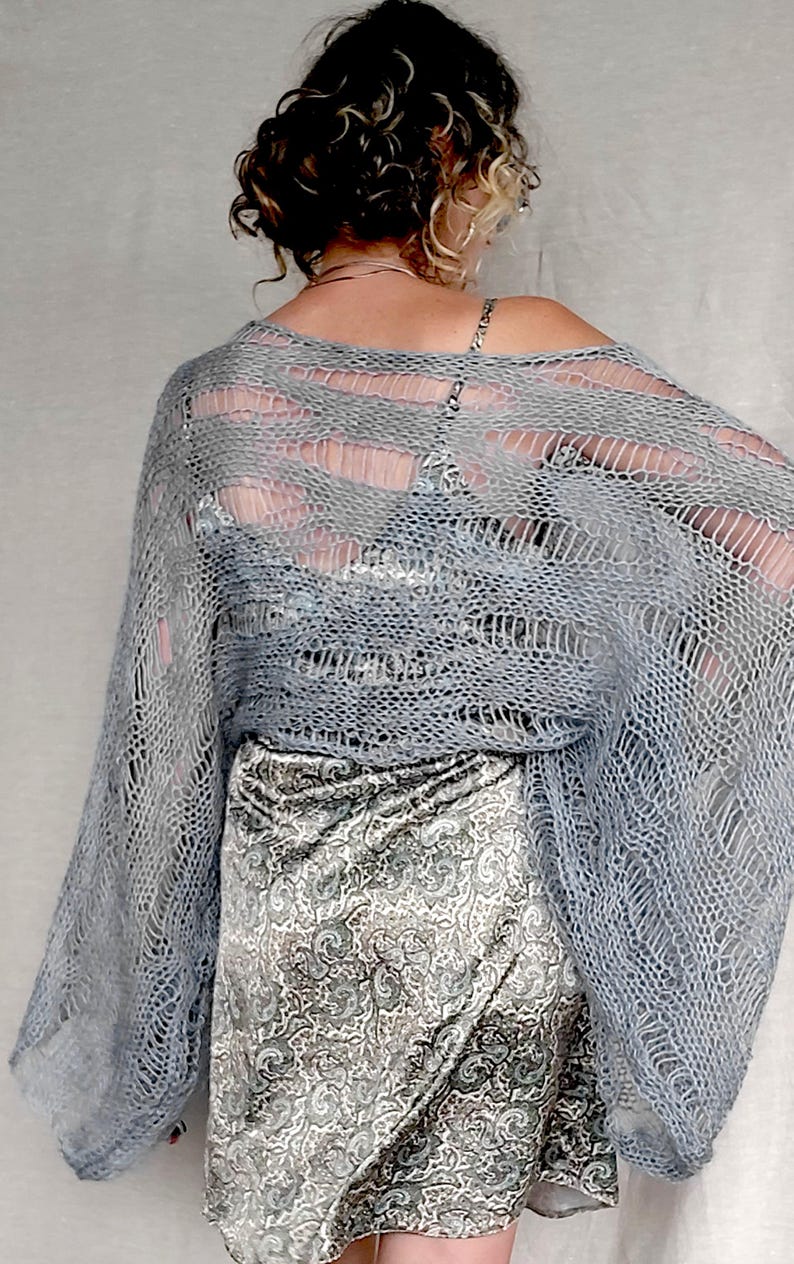 Op de afbeelding: Een lichtblauwe, open geweven gebreide sjaal met lange mouwen. De sjaal is over een rok met patroon gedrapeerd. Het breisel heeft een los, luchtig ontwerp. De rok heeft een paisley patroon in grijstinten en wit.