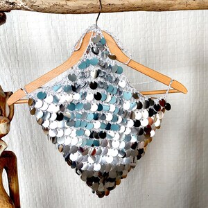 Eye-catching silver sequin top in a halter style, made with open-knit crochet and layers of round metallic discs. The pointed hemline and bold shine give it a fierce, futuristic edge. Perfect for dancers, performers, or daring fashion lovers.