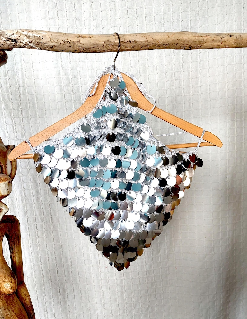 Eye-catching silver sequin top in a halter style, made with open-knit crochet and layers of round metallic discs. The pointed hemline and bold shine give it a fierce, futuristic edge. Perfect for dancers, performers, or daring fashion lovers.