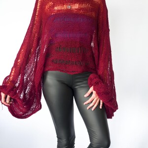 A burgundy knit poncho with wide sleeves. The open weave top is paired with dark gray leggings. The model is wearing sunglasses and a necklace. The background is white.