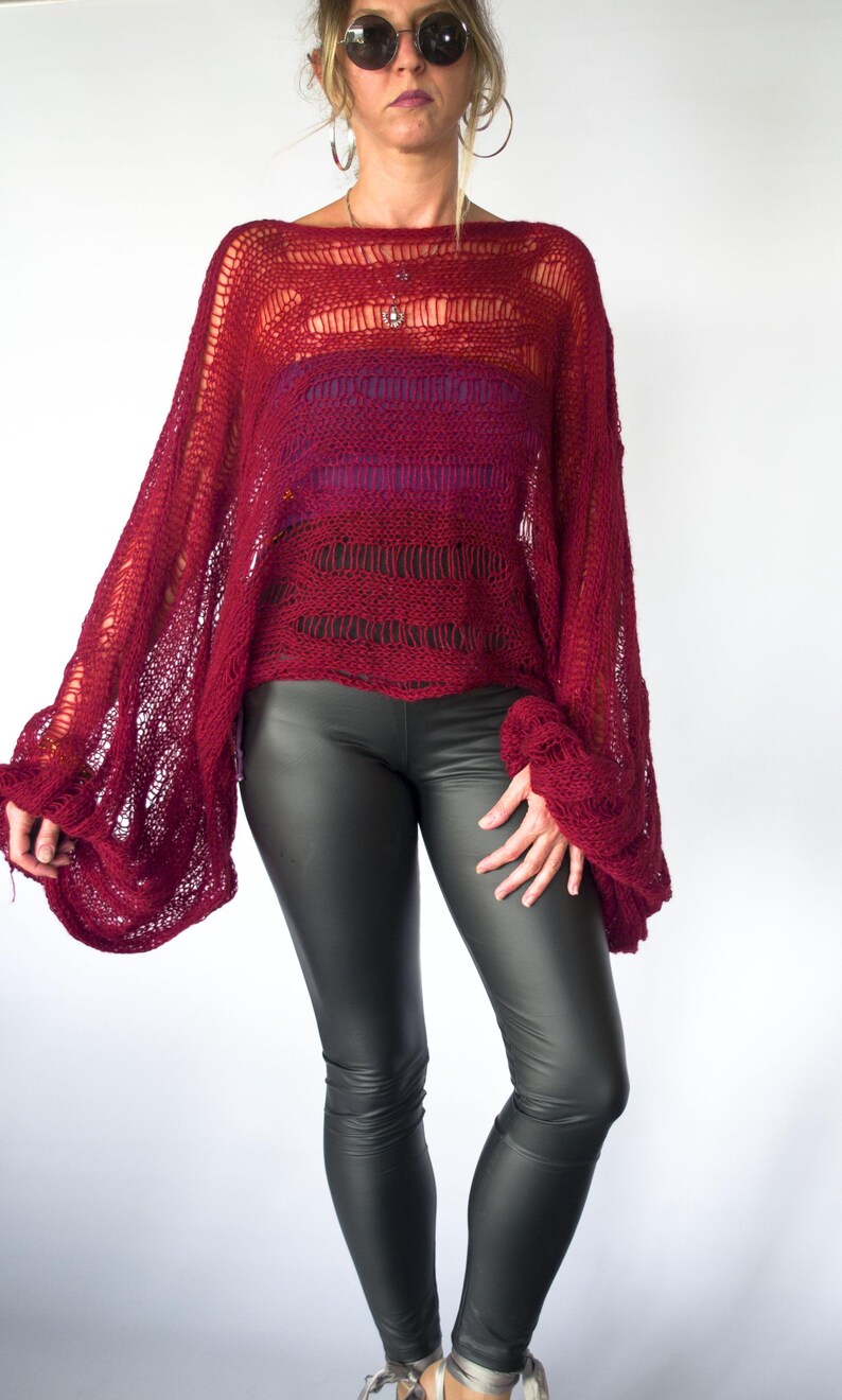 A burgundy knit poncho with wide sleeves. The open weave top is paired with dark gray leggings. The model is wearing sunglasses and a necklace. The background is white.