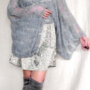 Op de afbeelding: Een lichtgrijze, open geweven gebreide sjaal met lange mouwen. De sjaal is gedrapeerd over een patroonjurk en gecombineerd met grijze dijhoge laarzen. De outfit is een mix van texturen en tinten, waardoor een zachte, gelaagde look ontstaat.