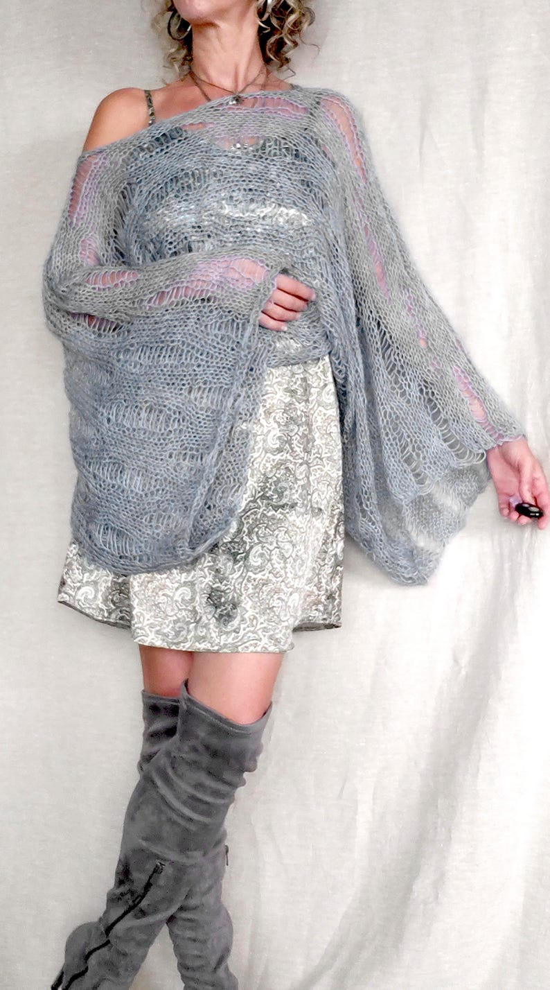 Op de afbeelding: Een lichtgrijze, open geweven gebreide sjaal met lange mouwen. De sjaal is gedrapeerd over een patroonjurk en gecombineerd met grijze dijhoge laarzen. De outfit is een mix van texturen en tinten, waardoor een zachte, gelaagde look ontstaat.