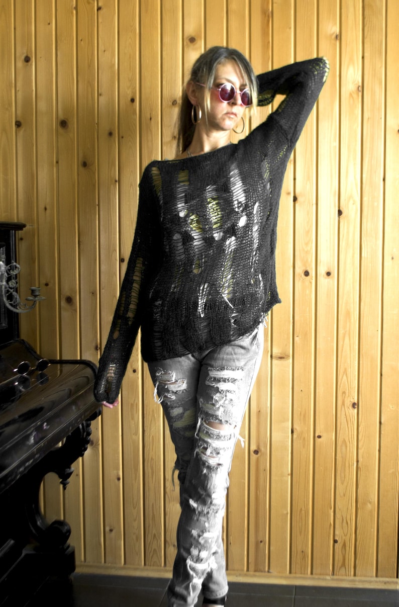 A black, open-weave sweater with distressed details is paired with ripped gray jeans. The outfit is worn by a person with pink sunglasses and hoop earrings. The background is a wooden wall.
