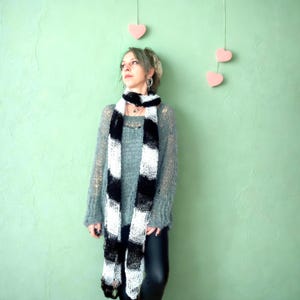 Könnte beinhalten: Ein schwarz-weiß gestreifter Schal ist über einem grauen Pullover drapiert. Der Schal hat ein kühnes, grafisches Design. Der Pullover ist ein locker gestrickter Stil. Der Hintergrund ist einfarbig hellgrün. Rosa Herzdekorationen hängen von oben.