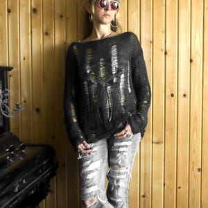 Black open-weave, heavily distressed knit sweater paired with ripped gray jeans; model with pink sunglasses and hoop earrings, wooden wall backdrop