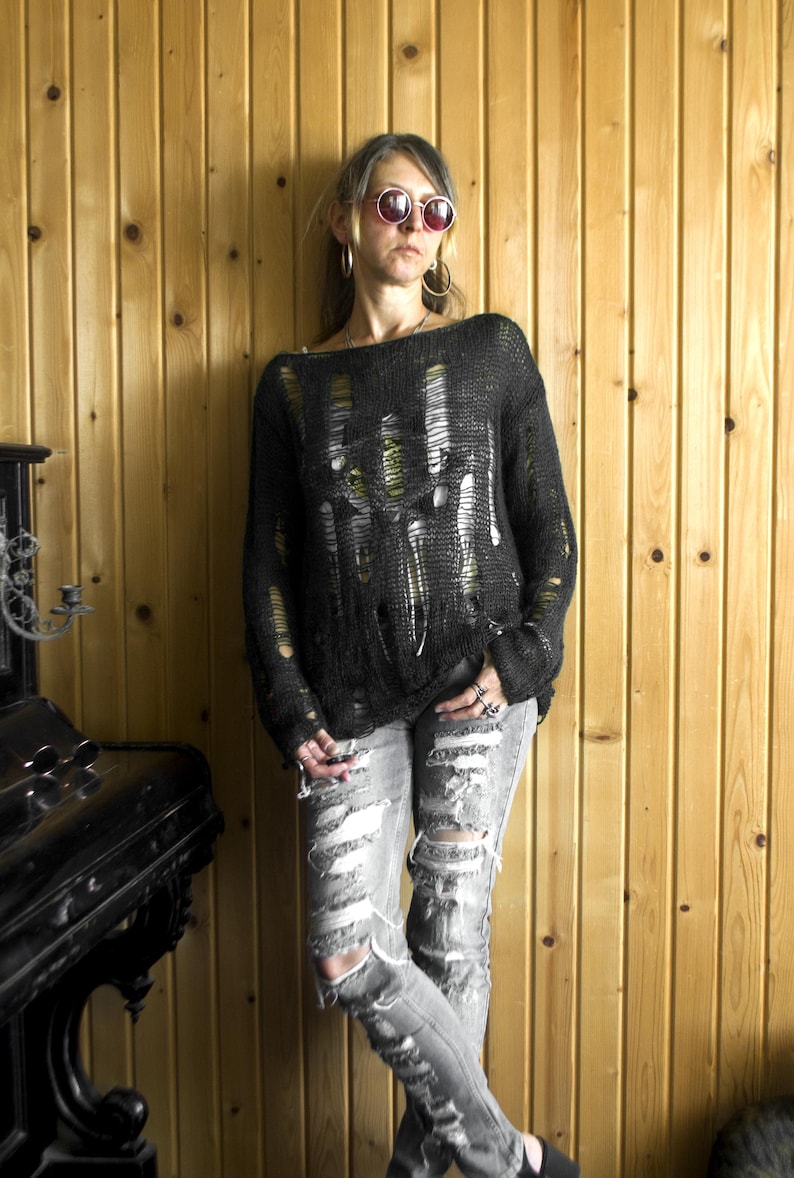 Black open-weave, heavily distressed knit sweater paired with ripped gray jeans; model with pink sunglasses and hoop earrings, wooden wall backdrop